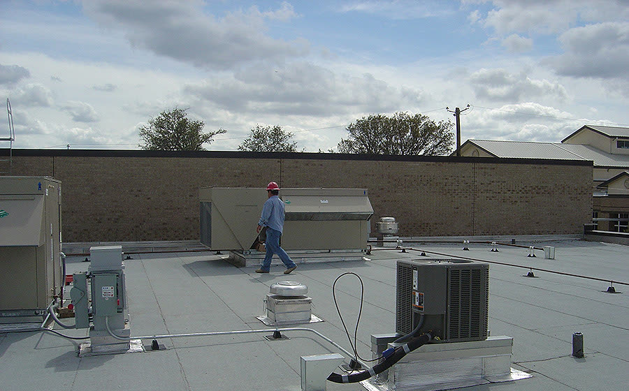 A worker in a red hard hat walking past a rooftop unit and a condensing unit, from Rabroker’s project gallery